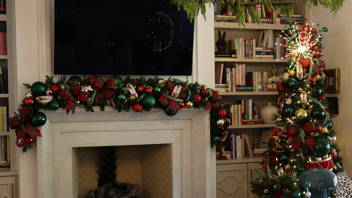 A cozy holiday living room decorated with a green wreath chandelier, a fireplace mantel garland, a decorated Christmas tree, bookshelves, and white sofas with patterned pillows.