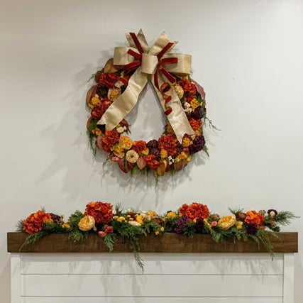 A decorative autumn wreath with a large cream ribbon bow hangs on a white wall above a mantel decorated with matching fall floral garlands.