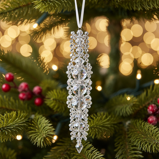 A long, sparkling silver Christmas tree ornament with clear crystal-like details hangs from a green pine tree branch with warm bokeh lights and red berries in the background.