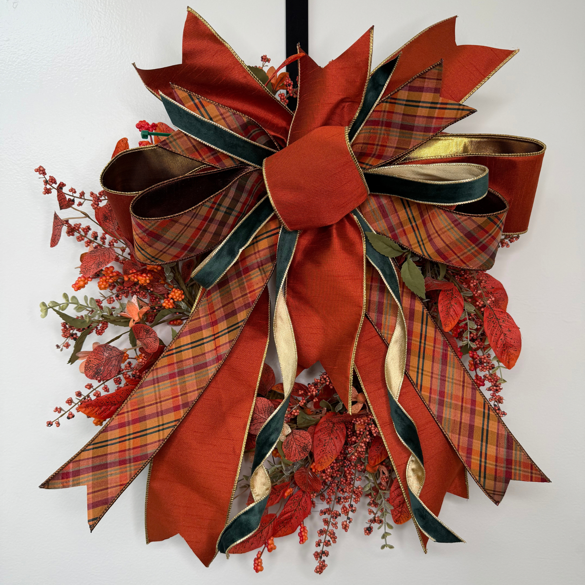 A large autumn-themed decorative bow wreath with layered ribbons in rust red, gold, and dark green plaid, accented with red berries and faux fall leaves on a white wall.