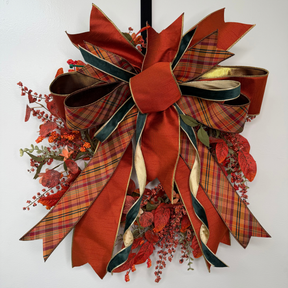 A large autumn-themed decorative bow wreath with layered ribbons in rust red, gold, and dark green plaid, accented with red berries and faux fall leaves on a white wall.