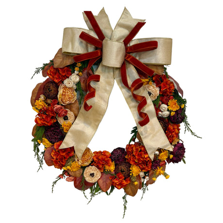 An autumn floral wreath decorated with orange, red, yellow, and brown faux flowers, green foliage, and a large cream satin bow with dark red ribbon accents, shown on a plain white background.