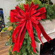 A large red velvet Christmas tree bow with long ribbon tails, attached to a green artificial pine garland in a white fireplace mantel setting.