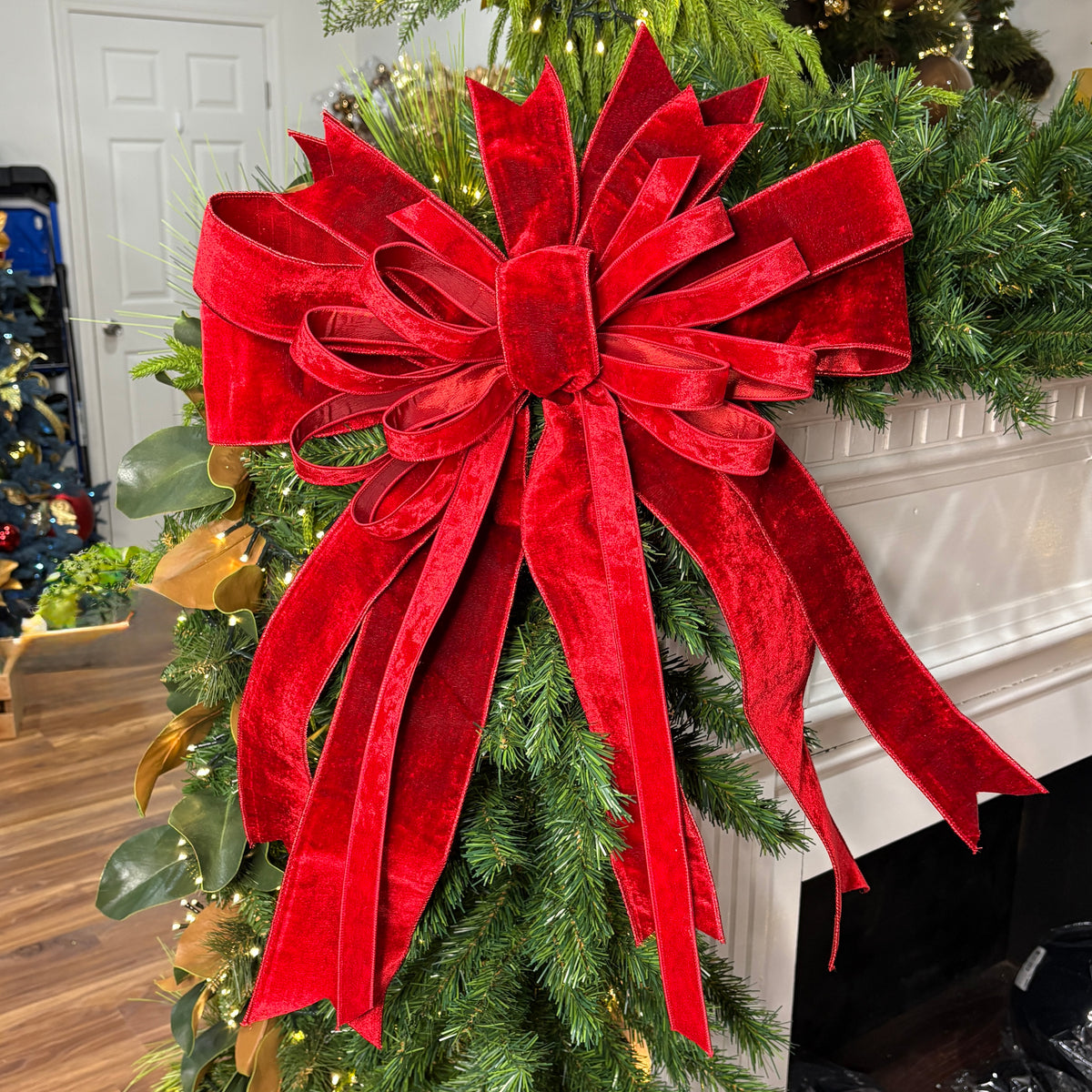 A large red velvet Christmas tree bow with long ribbon tails, attached to a green artificial pine garland in a white fireplace mantel setting.