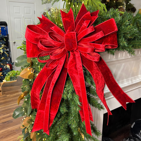 A large red velvet Christmas tree bow with long ribbon tails, attached to a green artificial pine garland in a white fireplace mantel setting.