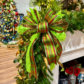 A large Christmas bow made from green and red plaid ribbon with glitter trim, tied around a green evergreen garland in front of a white fireplace mantel. A decorated Christmas tree and holiday decor are visible in the background.