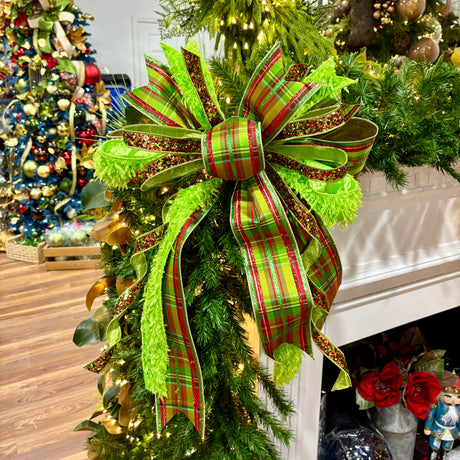 A large Christmas bow made from green and red plaid ribbon with glitter trim, tied around a green evergreen garland in front of a white fireplace mantel. A decorated Christmas tree and holiday decor are visible in the background.