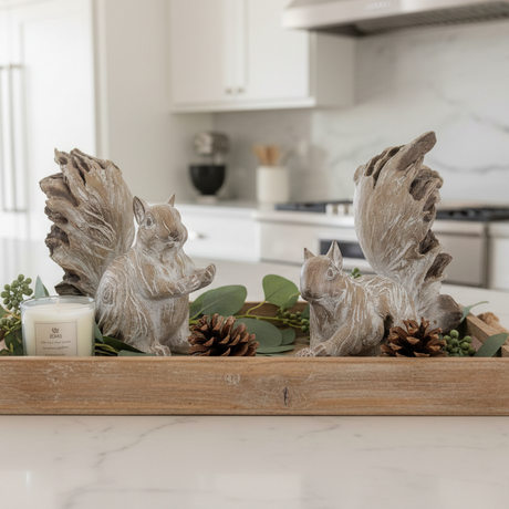 A rustic wooden tray centerpiece featuring two gray squirrel figurines, green eucalyptus leaves, pinecones, and a small white candle, displayed on a white marble kitchen countertop.