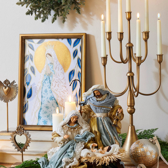 A styled Christmas tabletop scene with a nativity set in the center, a large gold candelabra with lit candles on the right, a framed religious painting on the left, and greenery and ornaments around it.