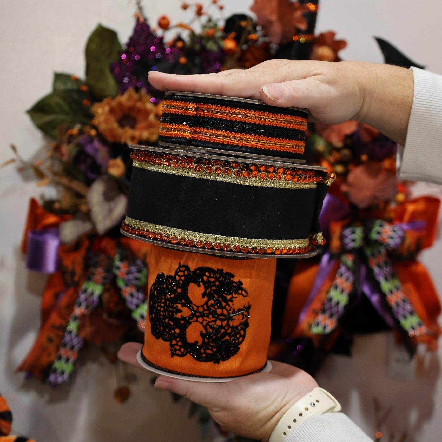 A person holds a stack of three orange and black Halloween-themed decorative ribbons s in front of a busy autumn wreath display.
