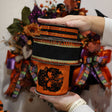 A person holds a stack of three orange and black Halloween-themed decorative ribbons s in front of a busy autumn wreath display.
