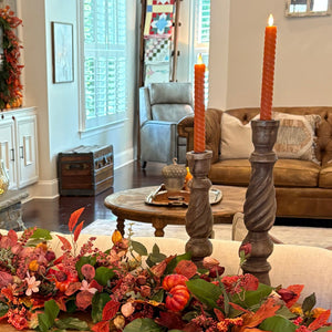 An autumn-themed console table centerpiece in a living room, featuring a floral garland with orange and red berries, green leaves, three orange pumpkins, and two tall candlesticks with orange taper candles.