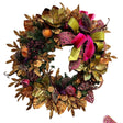An autumn-themed artificial wreath decorated with metallic gold and bronze leaves, green pine branches, dried orange slices, purple berries, and a large pink and lime green ribbon bow, shown against a plain off-white wall. Christmas wreath in a non-traditional color scheme. Falliday