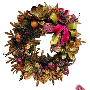 An autumn-themed artificial wreath decorated with metallic gold and bronze leaves, green pine branches, dried orange slices, purple berries, and a large pink and lime green ribbon bow, shown against a plain off-white wall. Christmas wreath in a non-traditional color scheme. Falliday
