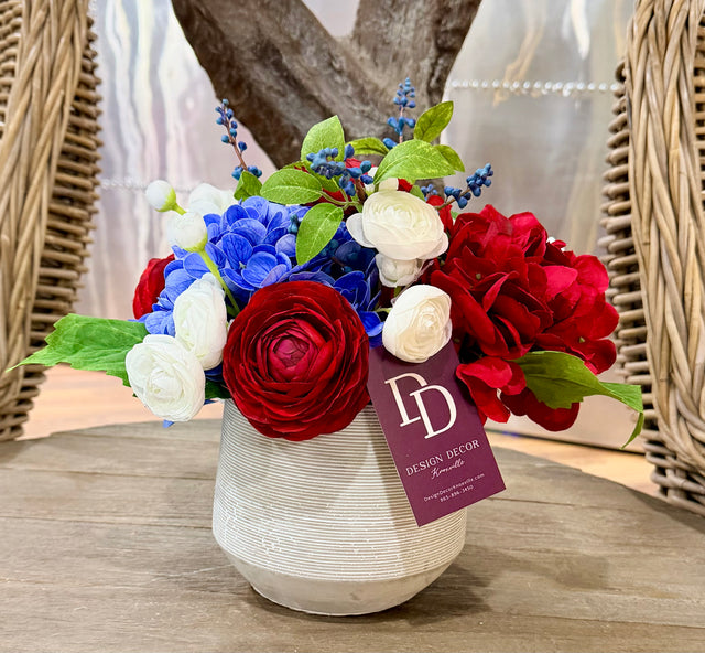 Floral arrangement with red, white, and blue flowers in a white vase on a wooden surface, with a 'Design Deco' tag.