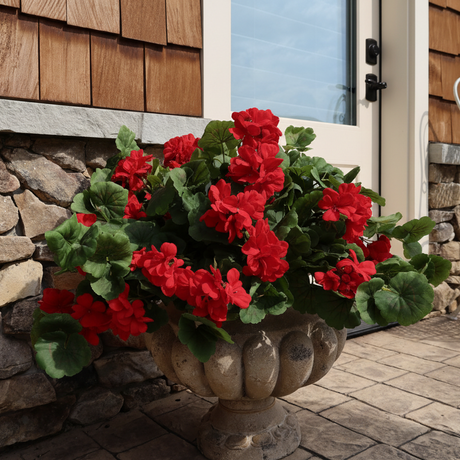 Red flowers in a stone planter on a patio with a wooden wall and white chair in the background.