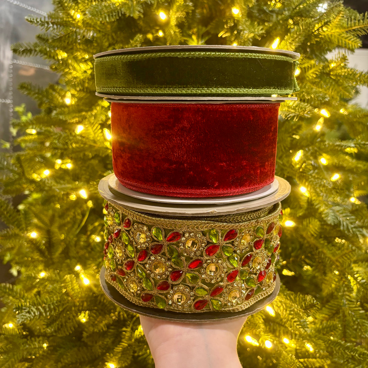 Three stacked rolls of decorative ribbon held by a hand in front of a glowing Christmas tree. The top roll is olive green velvet, the middle is red velvet, and the bottom is a wide ornate gold ribbon embellished with red and green faux jewels.