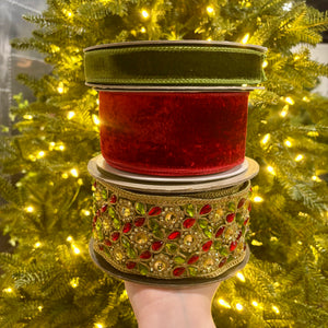 Three stacked rolls of decorative ribbon held by a hand in front of a glowing Christmas tree. The top roll is olive green velvet, the middle is red velvet, and the bottom is a wide ornate gold ribbon embellished with red and green faux jewels.