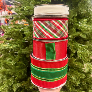 A stack of four Christmas-themed grosgrain ribbon spools held in front of a green Christmas tree. The ribbons feature red, green, and white patterns including plaid, stripes, and a solid green accent.