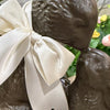 Faux chocolate sculpture of two kissing bunnies with a large white bow, set against a blurred floral background.