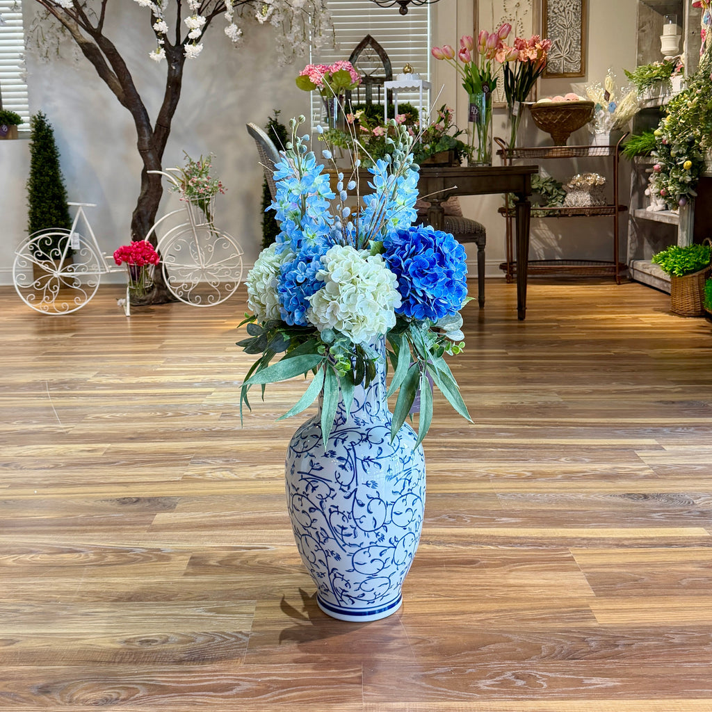 Decorative floral arrangement in a blue and white vase on a wooden floor with a background of indoor plants and decor.