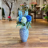 Decorative floral arrangement in a blue and white vase on a wooden floor with a background of indoor plants and decor.
