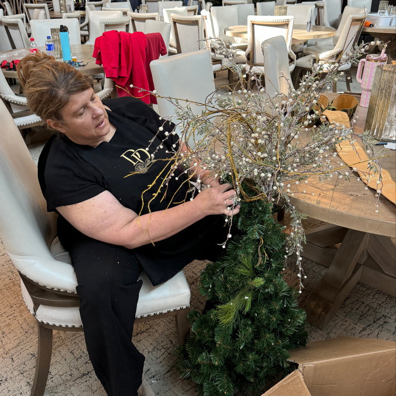 A woman sits in a dining room setting, assembling a decorative holiday tree with green pine branches, silver bead garlands, and gold wire accents.