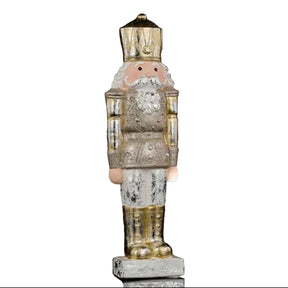 A vintage-style nutcracker figurine with a distressed silver and gold finish, featuring a pink face, white beard, and standing on a small base against a black background.