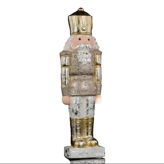 A vintage-style nutcracker figurine with a distressed silver and gold finish, featuring a pink face, white beard, and standing on a small base against a black background.