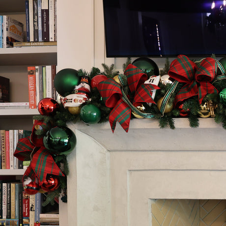 A festive Christmas garland decorated with green pine branches, red and green ornaments, and large red plaid bows, draped over a white fireplace mantel in a home interior setting.